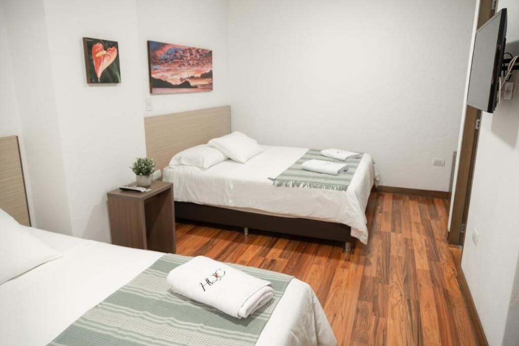 A hotel room with two beds, a wooden floor, a TV mounted on the wall, and two paintings on the wall.