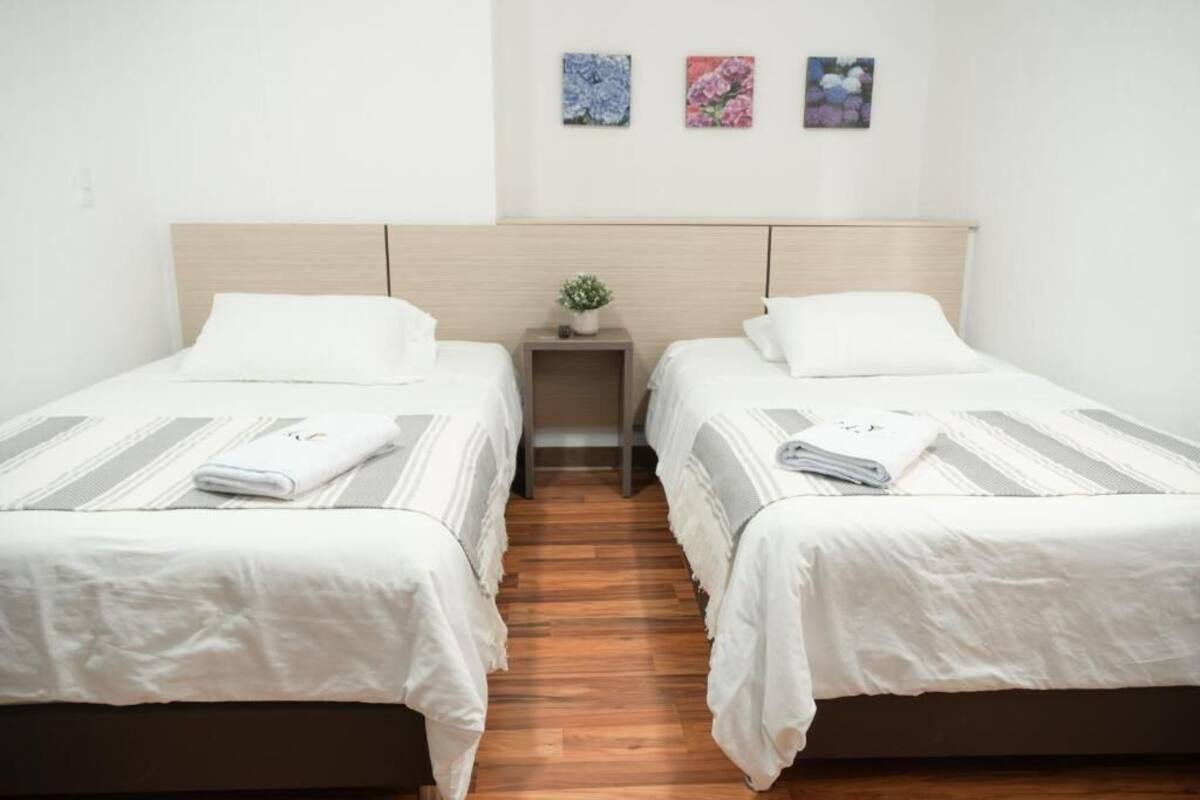 A hotel room with two single beds, a wooden headboard, a small bedside table with a plant, and three framed floral artworks on the wall.