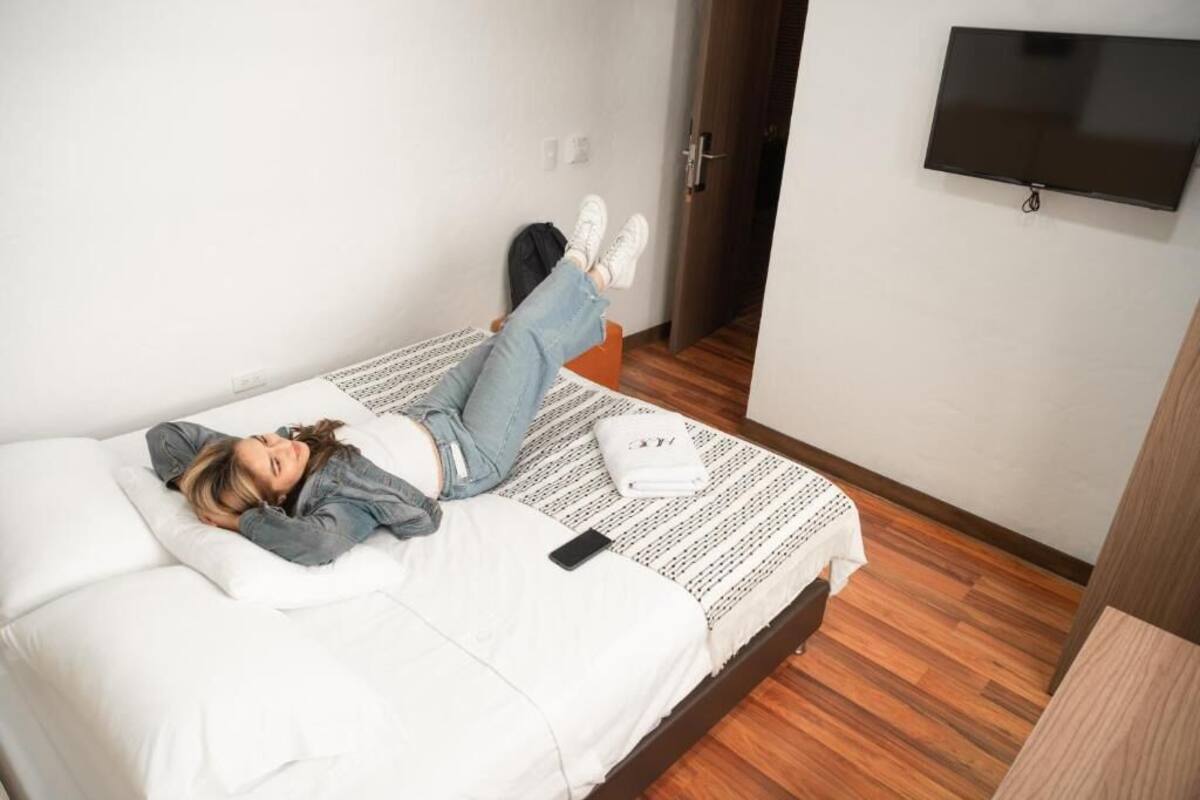 A person lying on a bed in a room with a wooden floor, a white wall, and a flat-screen TV mounted on the wall.