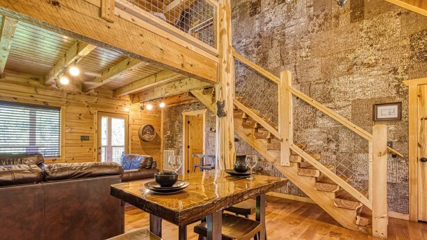 Ein rustikaler Innenraum mit einer Holztreppe, einem Esstisch mit Hockern, einem Ledersofa und einem Kamin.