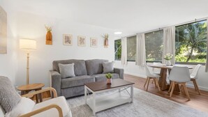Uma sala de estar moderna com um sofá cinzento, uma mesa de centro e um espaço de jantar com mesa e cadeiras. Há quadros nas paredes e um abajur de chão.