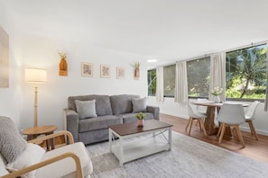 Un salón moderno con un sofá gris, una mesa de centro y un comedor con mesa y sillas. Hay cuadros enmarcados en la pared y una lámpara de pie.