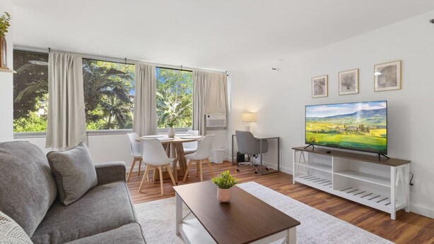 Uma sala de estar moderna com um sofá, uma mesa de jantar com cadeiras, uma televisão sobre um móvel e janelas amplas com cortinas.