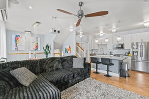 A modern kitchen with stainless steel appliances, a bar area, and a large black sectional sofa. - Modern Island Living | Parking, AC, Fast WiFi, W/D (Ewa Beach)