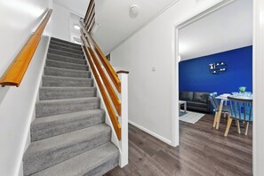 Un escalier avec des rampes en bois mène à une pièce dont un des murs est peint en bleu. On y trouve également un coin repas avec une table et des chaises, ainsi qu’un canapé.