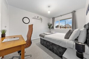 Une chambre moderne avec un lit, un bureau et une chaise. Une fenêtre offre une vue sur les maisons à l’extérieur.