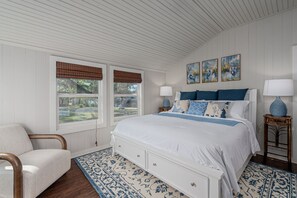 A bedroom with a large bed, two bedside tables with lamps, a chair, and a window with blinds.