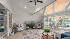 Sala de estar ampla com uma TV grande, assentos confortáveis, ventilador de teto e vista para o exterior.