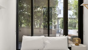 A bedroom with a bed, white pillows, a bedside table with a lamp, and a view of trees and a balcony. - Bright Inner-north Stay With Pool Access (Melbourne)