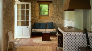 Une pièce rustique aux murs de pierre, avec un banc en bois agrémenté de coussins, une porte blanche et une fenêtre donnant sur un paysage verdoyant.