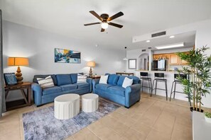 A modern living room with a blue sofa set, a ceiling fan, and a kitchen area with bar stools. - Nice cozy Lake condo, Disney & NASA launches view! (Orlando)