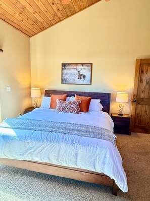A bedroom with a wooden ceiling, a bed with patterned bedding, two bedside lamps, a framed picture on the wall, and a wooden door. - Rim Retreat in the Pines, modern, cozy cabin nestled in the Ponderosa Pines. (Pine)