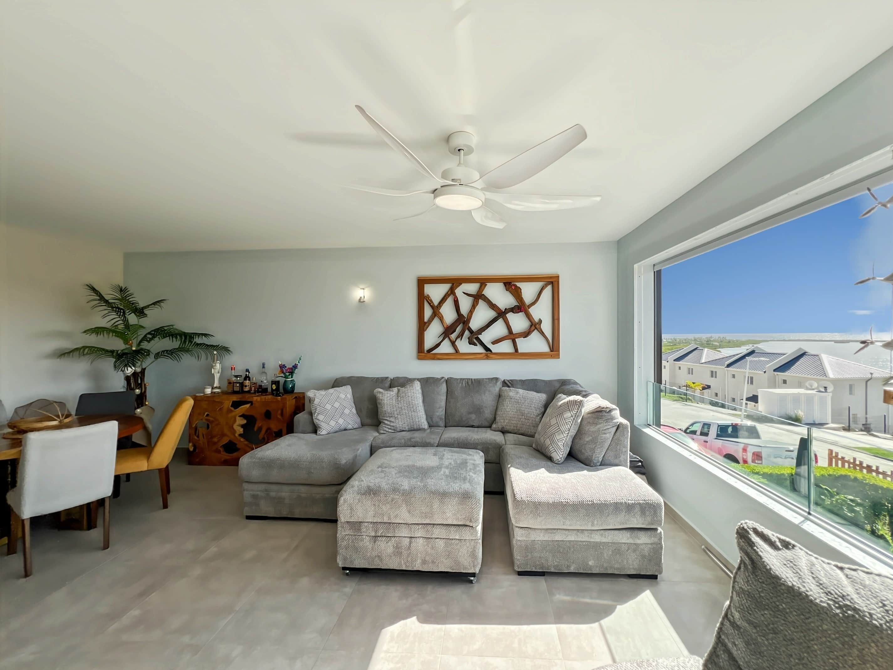 Uma sala de estar moderna com sofá seccional, pufes, área de jantar com mesa e cadeiras, ventilador de teto e uma grande janela com vista para casas.
