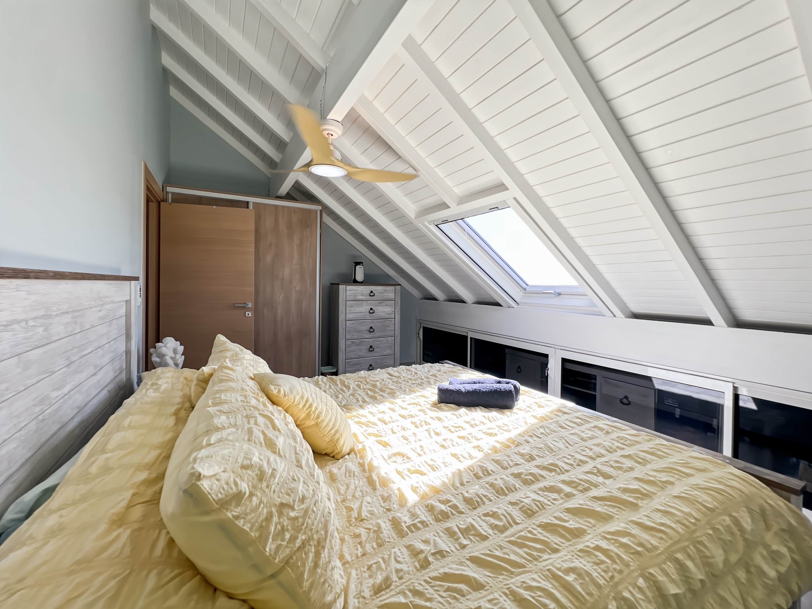A bedroom with a sloped ceiling, a bed with a textured duvet, a wooden wardrobe, and a desk with drawers.