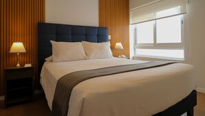 Hotellihuone, jossa on suuri sänky, kaksi yöpöytää, ikkuna, jossa on sälekaihtimet, ja yksi seinä on koristeltu puupaneloinnilla.