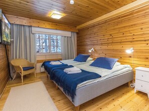 A wooden-paneled bedroom with a bed, two bedside tables, a chair, and a window with a view of snow-covered trees. - Saajomaja by Interhome (Inari)