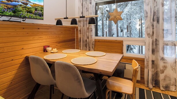 A dining area with a wooden table, four chairs, and a large window with a view of a snowy forest. - Villa rokua by Interhome (Utajärvi)