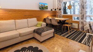 A cozy living area with a sofa, dining table, and chairs, wooden flooring, and a large window with curtains. - Villa rokua by Interhome (Utajärvi)