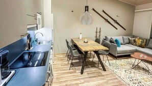 A modern kitchen with a built-in stove, a dining area with a wooden table and chairs, and a grey sofa in a living area with a patterned rug. - Pikkuhippu by Interhome (Pelkosenniemi)