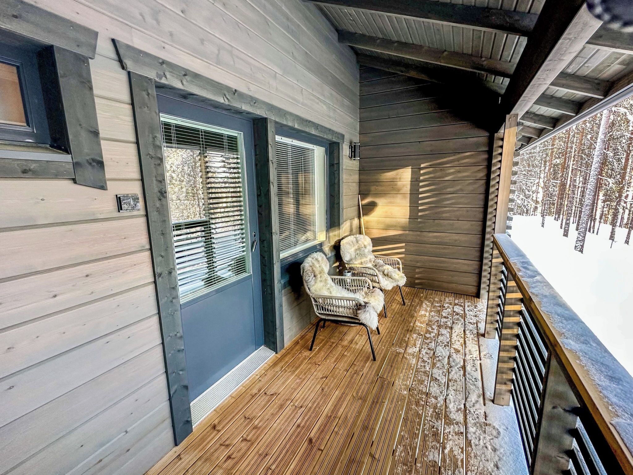 A wooden cabin with a covered porch, a dog sitting on a chair, and a snowy forest view.