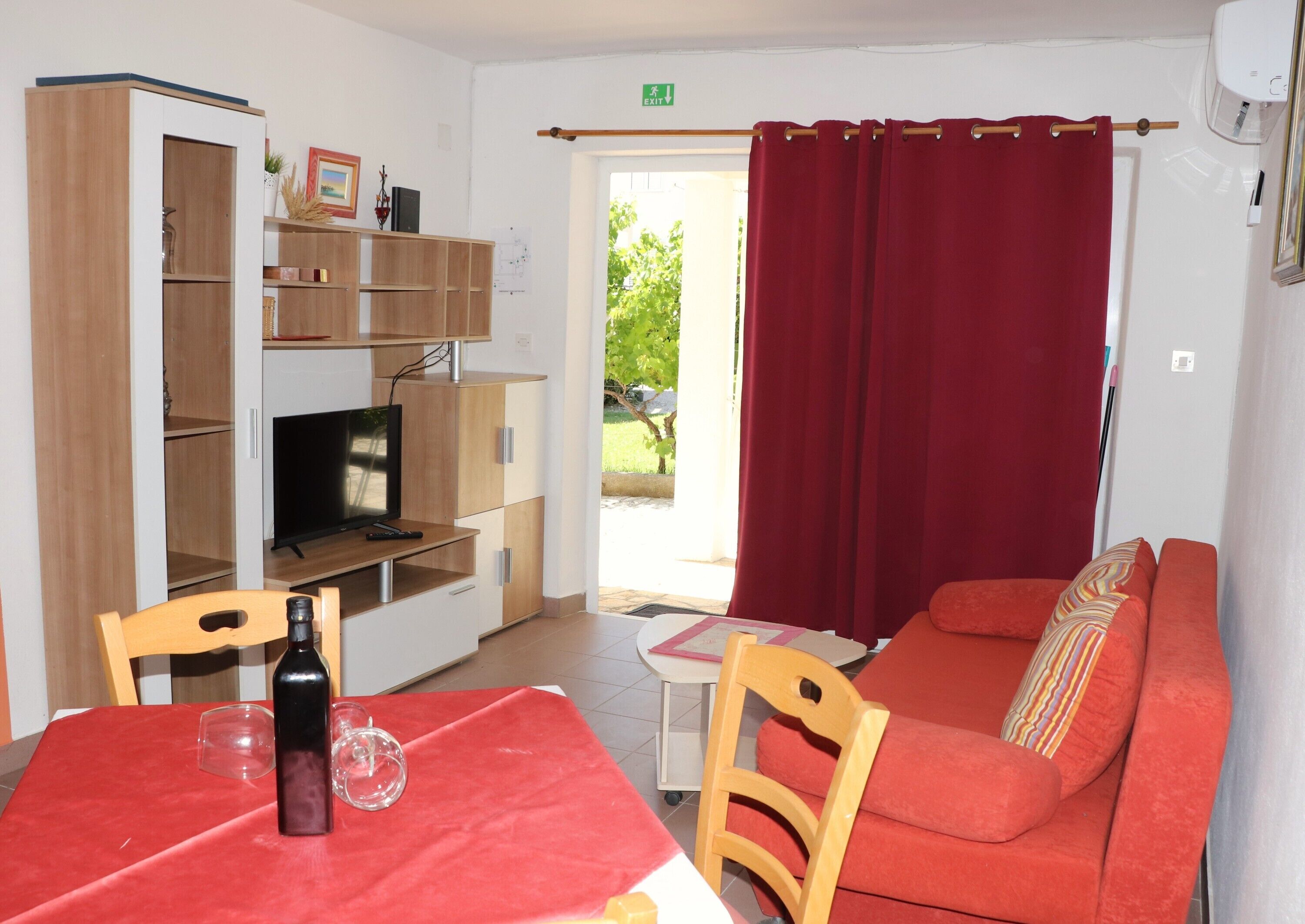 Un espace de vie compact comprenant un canapé, un téléviseur posé sur un meuble en bois, une table à manger avec une bouteille et un verre, et un rideau rouge.