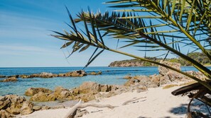 Uma praia rochosa com águas azuis cristalinas e uma colina distante com edifícios visíveis.