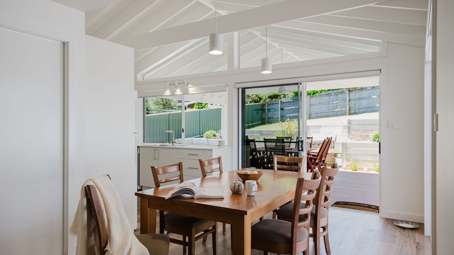 A modern kitchen with a large dining table, wooden chairs, and a view of the backyard through large windows.