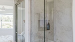 Een badkamer met een douchecabine van glas, een fles shampoo en een bed met witte lakens.