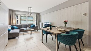 A modern living room with a sofa, a dining table with chairs, a small coffee table, and a window with curtains. - Walter Vilain 0301 + G* (Sint Idesbald)