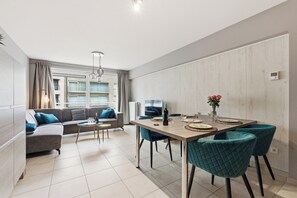 A modern living room with a sofa, a dining table with chairs, a small coffee table, and a window with curtains. - Walter Vilain 0301 + G* (Sint Idesbald)