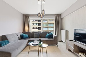 A modern living room with a grey sofa, a glass coffee table, a flat-screen TV, and a city view through large windows. - Walter Vilain 0301 + G* (Sint Idesbald)