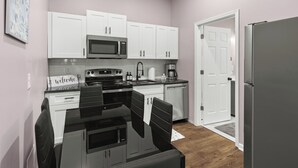 A compact kitchen with white cabinets, a microwave, a stove, and a refrigerator. There is a small dining area with a table and chairs. - Cozy Garden-Level Retreat 3 Queen Beds (Raleigh)