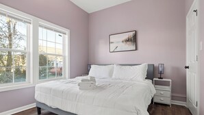 A bedroom with a large bed, a bedside table with a lamp, a window with blinds, and a door. - Cozy Garden-Level Retreat 3 Queen Beds (Raleigh)