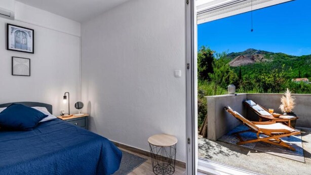 Un dormitorio con cama, mesita de noche, lámpara y una pequeña mesa redonda. Una puerta corrediza de vidrio da a un espacio exterior con una silla de descanso y una mesa.
