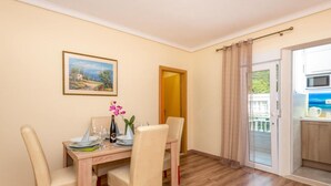 Un comedor con una mesa de madera dispuesta para cuatro, una botella de champán y un florero con flores rosas. Hay un cuadro en la pared y una puerta que da a un balcón con vistas al mar.