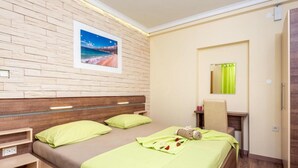 A hotel room with a wooden headboard, two beds with green bedding, a bedside table, a small desk with a chair, a mirror, and a picture of a beach on the wall. - Villa Goldfield - One Bedroom Apartment with Balcony (Gradac)