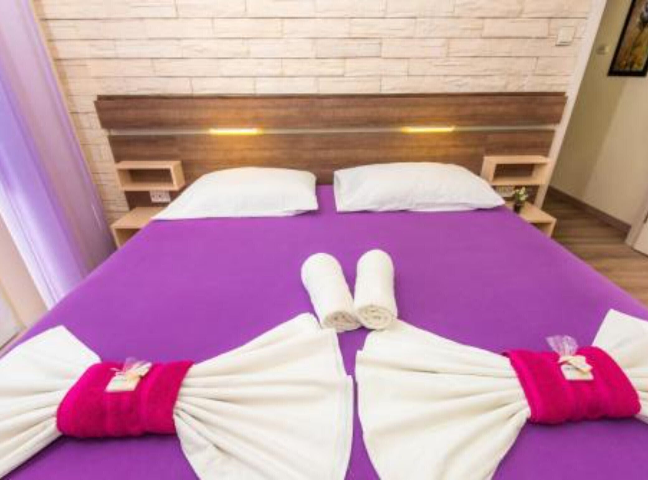 Ein Hotelzimmer mit violettem Bettbezug, einem Holzkopfteil, weißen Kissen und ordentlich drapierten Handtüchern auf dem Bett.