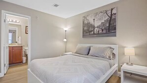 A bedroom with a large bed, a nightstand, a lamp, a painting, and a bathroom visible through an open door.