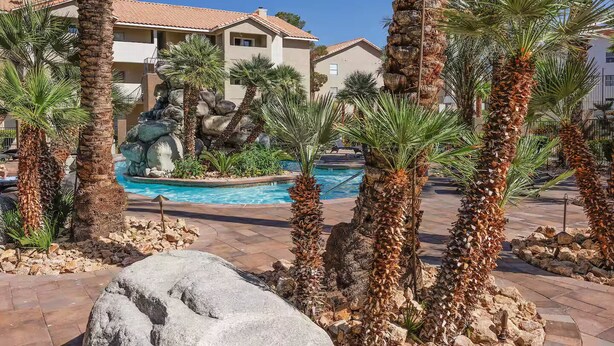 A pool area with palm trees and a rock formation, surrounded by buildings with a tiled roof.