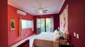 Ein Schlafzimmer mit einem Bett, einem Deckenventilator, einer Klimaanlage, einem Fenster mit Vorhängen und einer Wand mit Wandteppich.