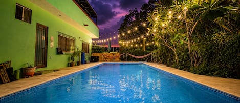 A swimming pool with a tiled edge, surrounded by greenery and a building with a green facade.