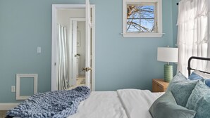 Ein Schlafzimmer mit Bett, Fenster, Nachttisch und einer Tür zu einem anderen Raum.