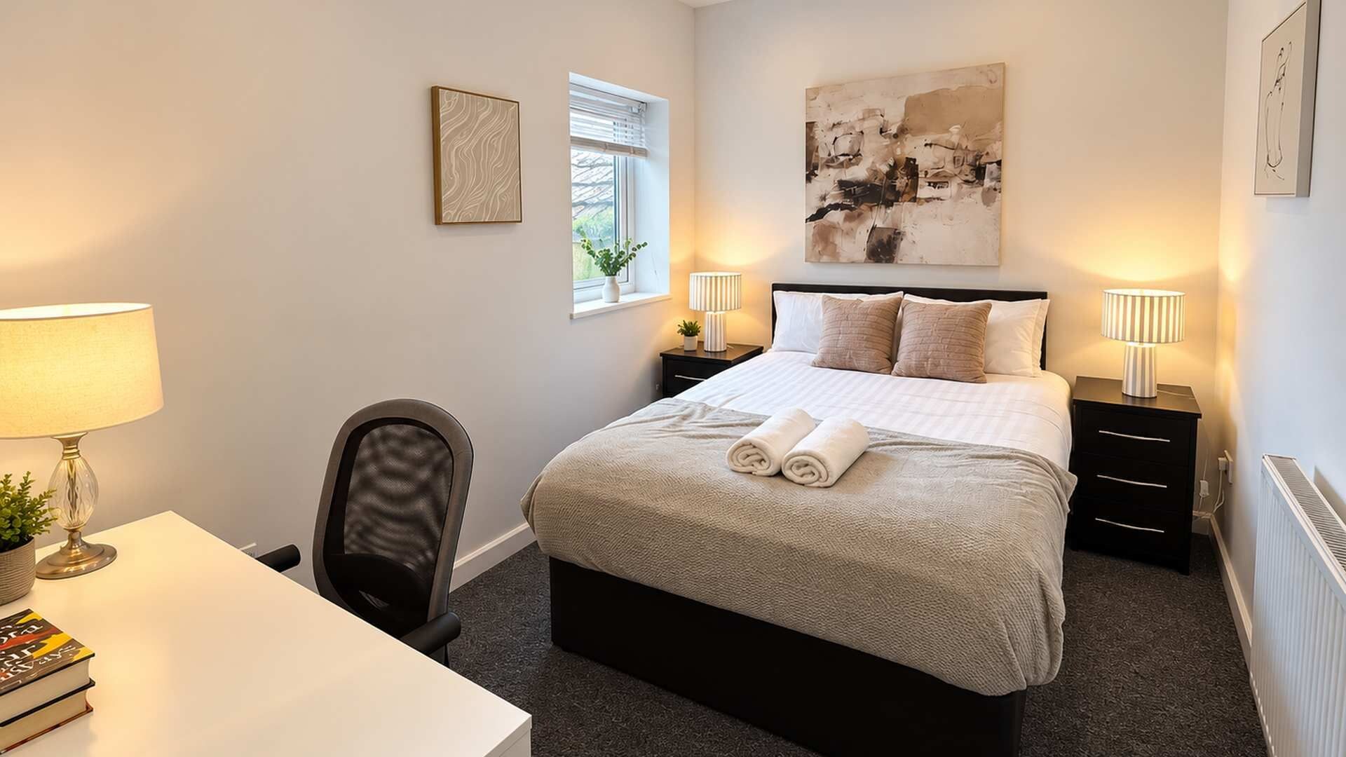 A hotel room with a bed, bedside tables, a desk, a chair, and artwork on the walls.