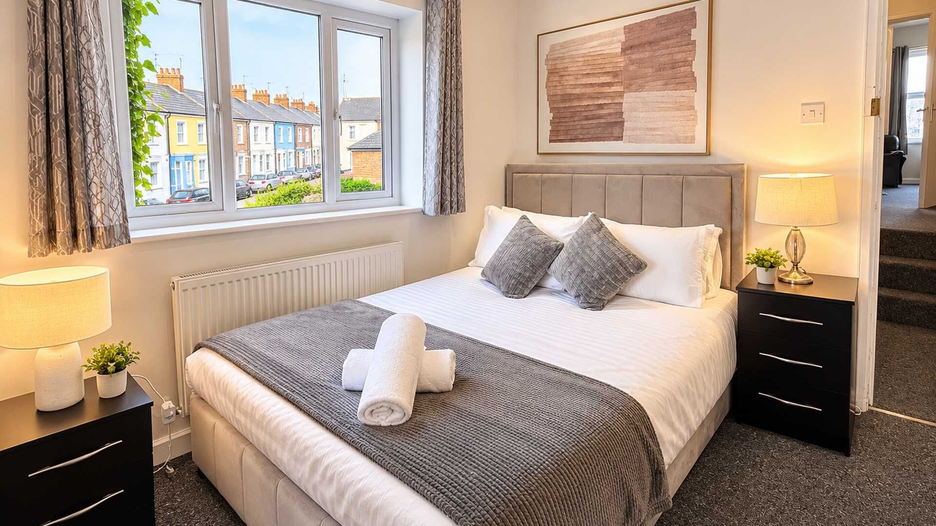 Een hotelkamer met een bed, nachtkastjes, een raam met gordijnen, een radiator, een lijst met een kunstwerk aan de muur en uitzicht op een woonstraat.
