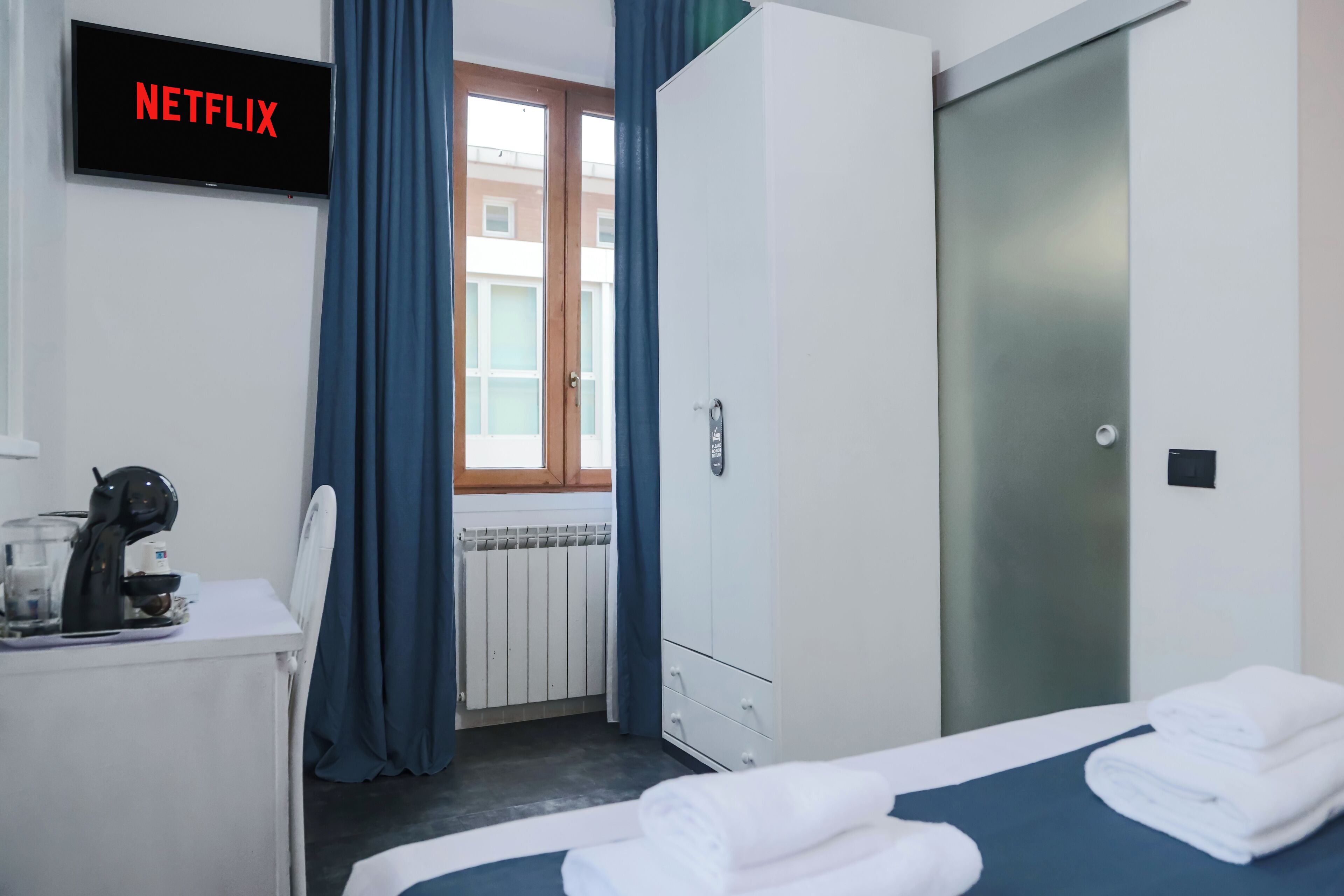 Une chambre d’hôtel avec un lit, un bureau doté d’une machine à café, une armoire et une vue sur l’extérieur par une fenêtre ornée de rideaux bleus.