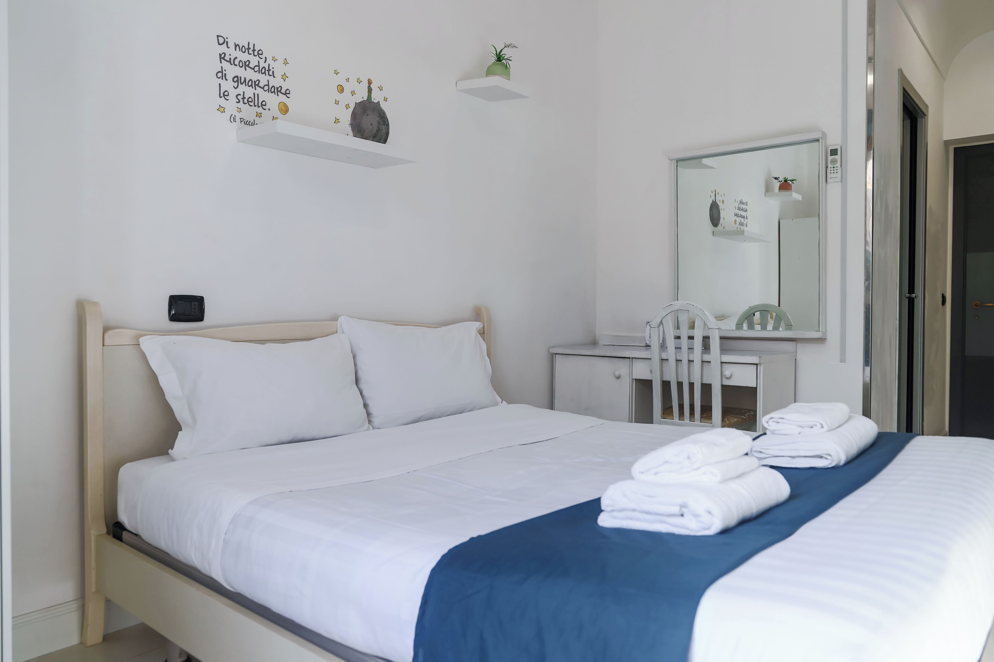 Ein ordentlich bezogenes Bett mit weißer und blauer Bettwäsche, ein Nachttisch mit Lampe, ein kleiner Schreibtisch mit Stuhl und ein Spiegel an der Wand.