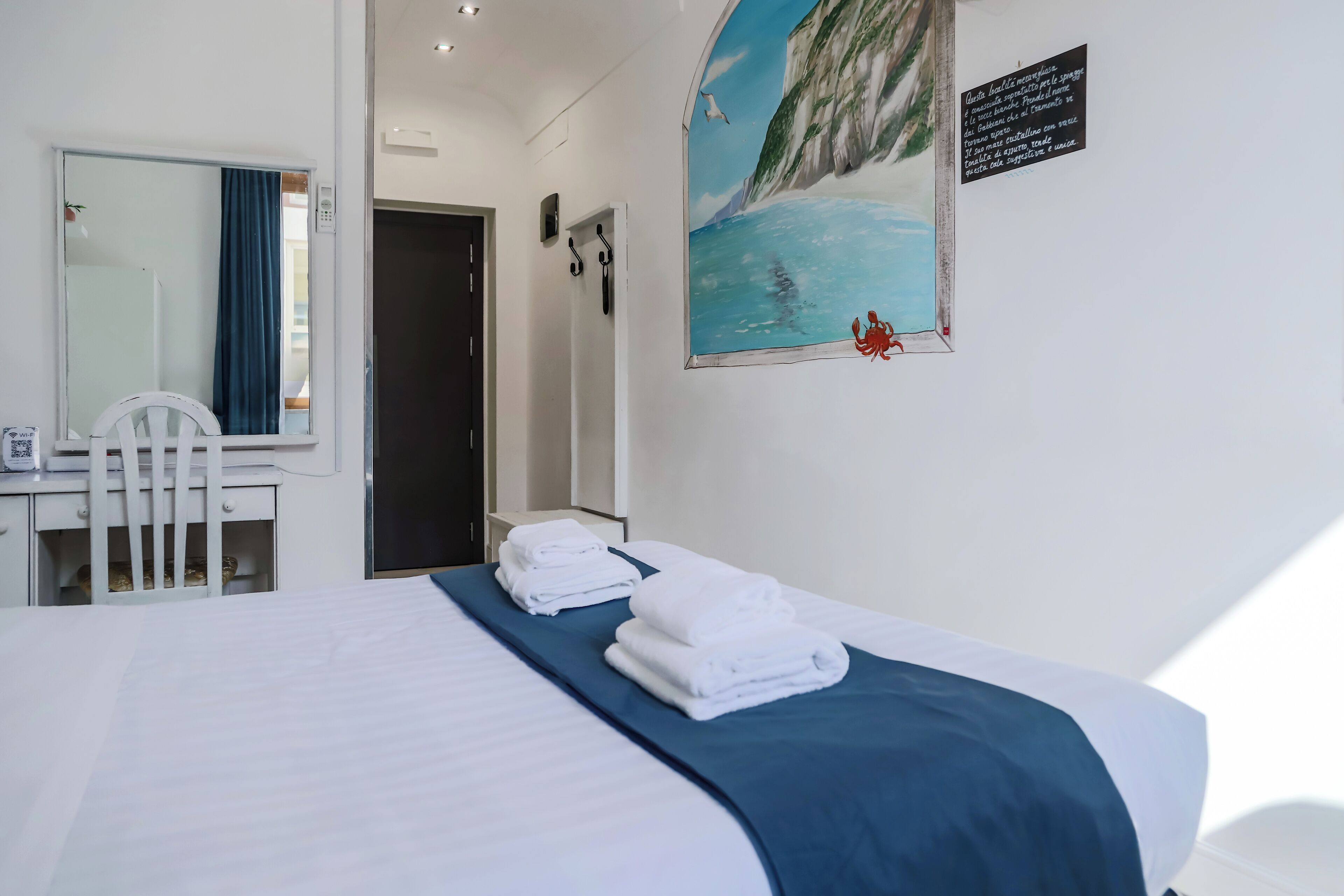 Hotelkamer met een bed, een bureau, een stoel, een spiegel en een schilderij van een kustlandschap.