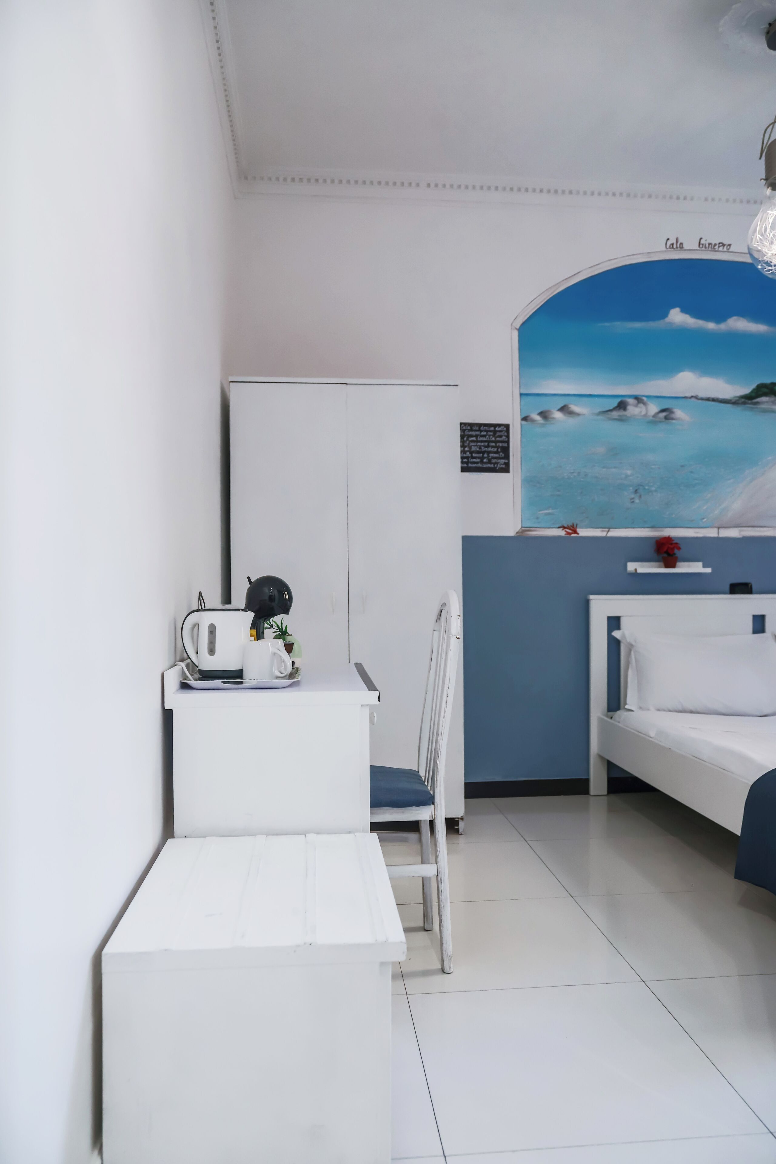 Een kleine, schone kamer met een bed, een stoel, een klein tafeltje met een waterkoker en een kop, en een schilderij van een strandlandschap aan de muur.
