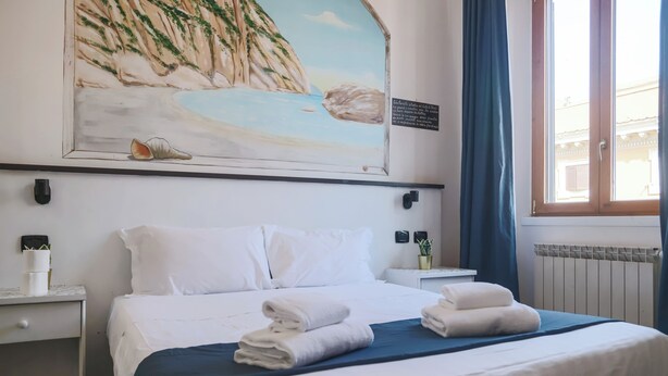 Hotellihuone, jossa on sänky, valkoiset vuodevaatteet, sininen peitto, rantamaiseman kuvaava taulu ja ikkuna, jossa on siniset verhot.