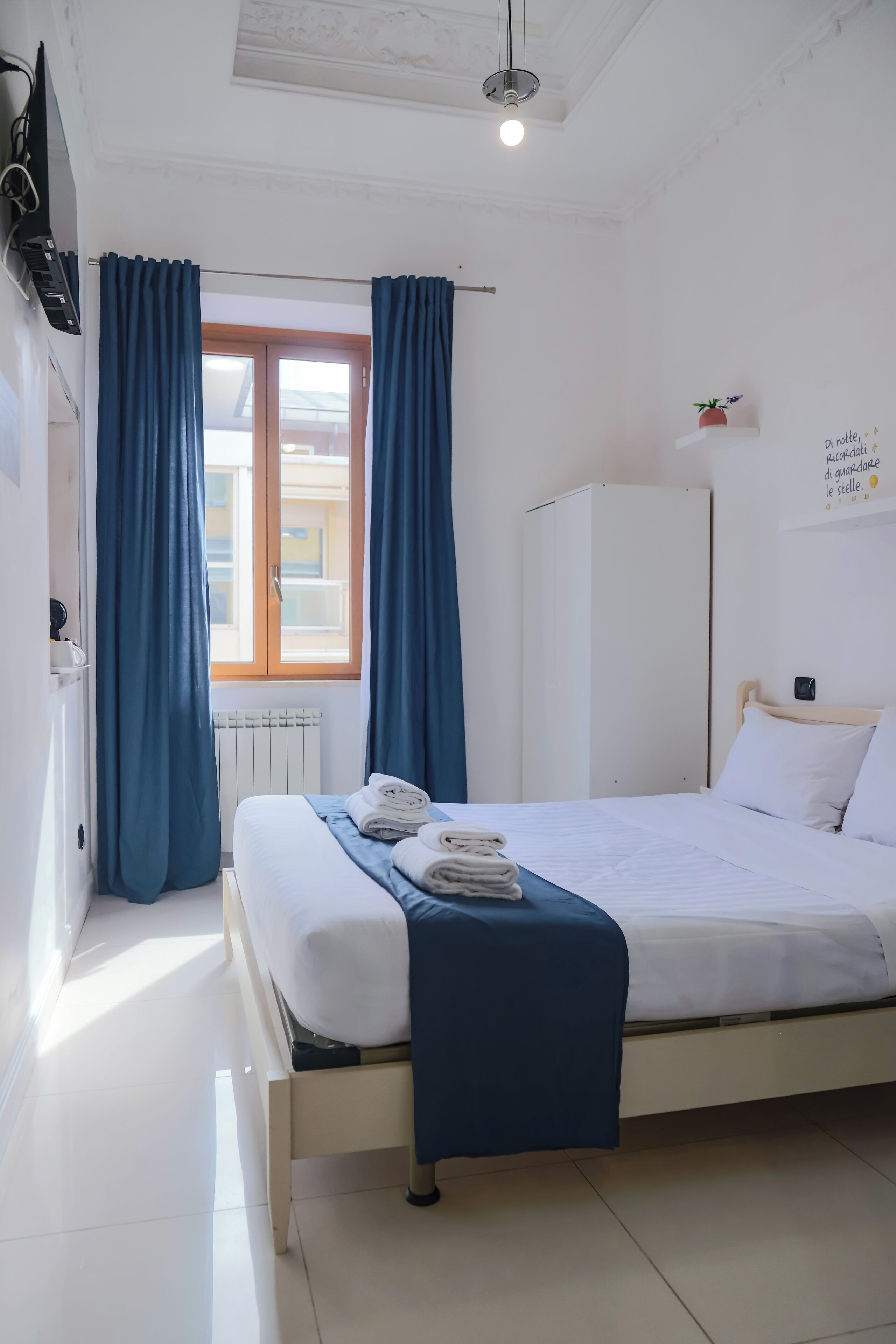A clean, well-lit hotel room with a bed, blue curtains, a radiator, and a window with a view of buildings.